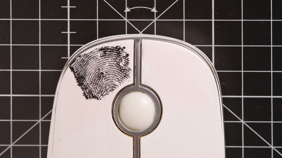 Mouse de computador com impressão digital destacada sob uma lente de aumento, sobre fundo técnico quadriculado.