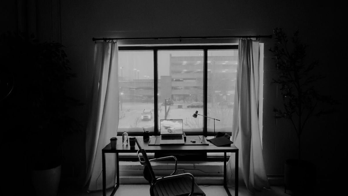 Escritório vazio com mesa, cadeira, computador portátil e janela ao fundo, em preto e branco.