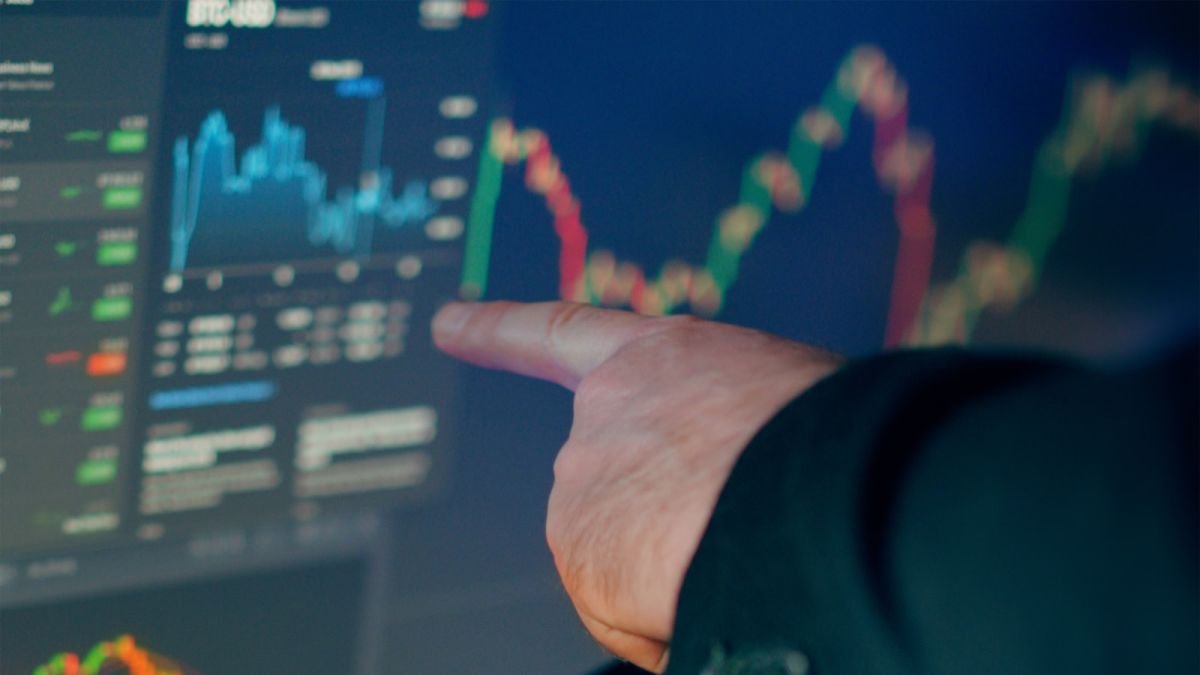 Profissional aponta para gráficos financeiros exibidos em várias telas digitais.