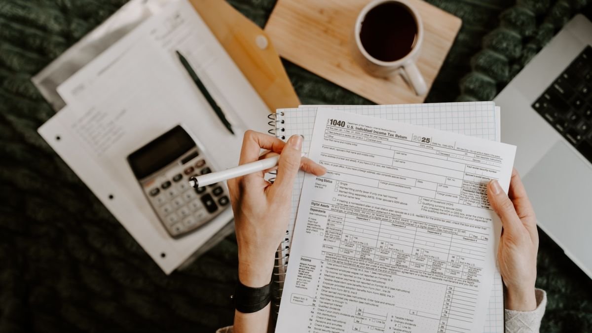 Pessoa revisa formulários com calculadora, caneta e computador portátil sobre uma mesa.